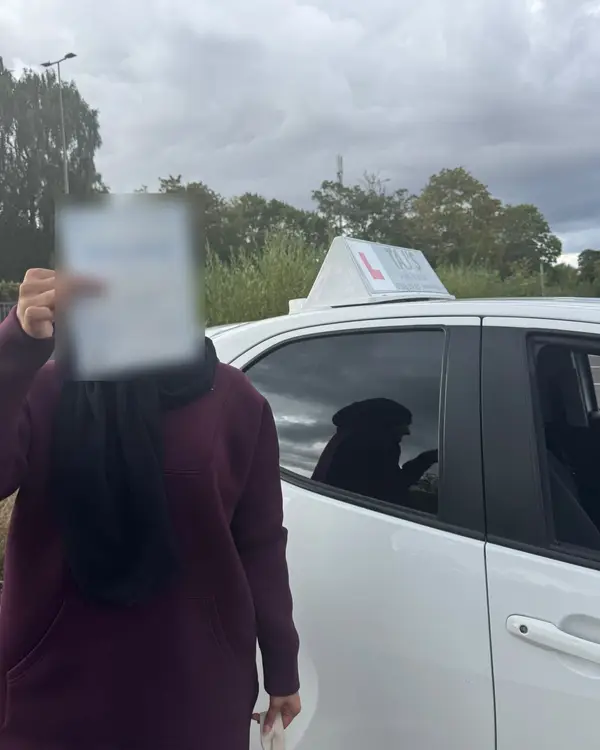 A learner holding their driving test pass certificate next to a Pass with Taj car